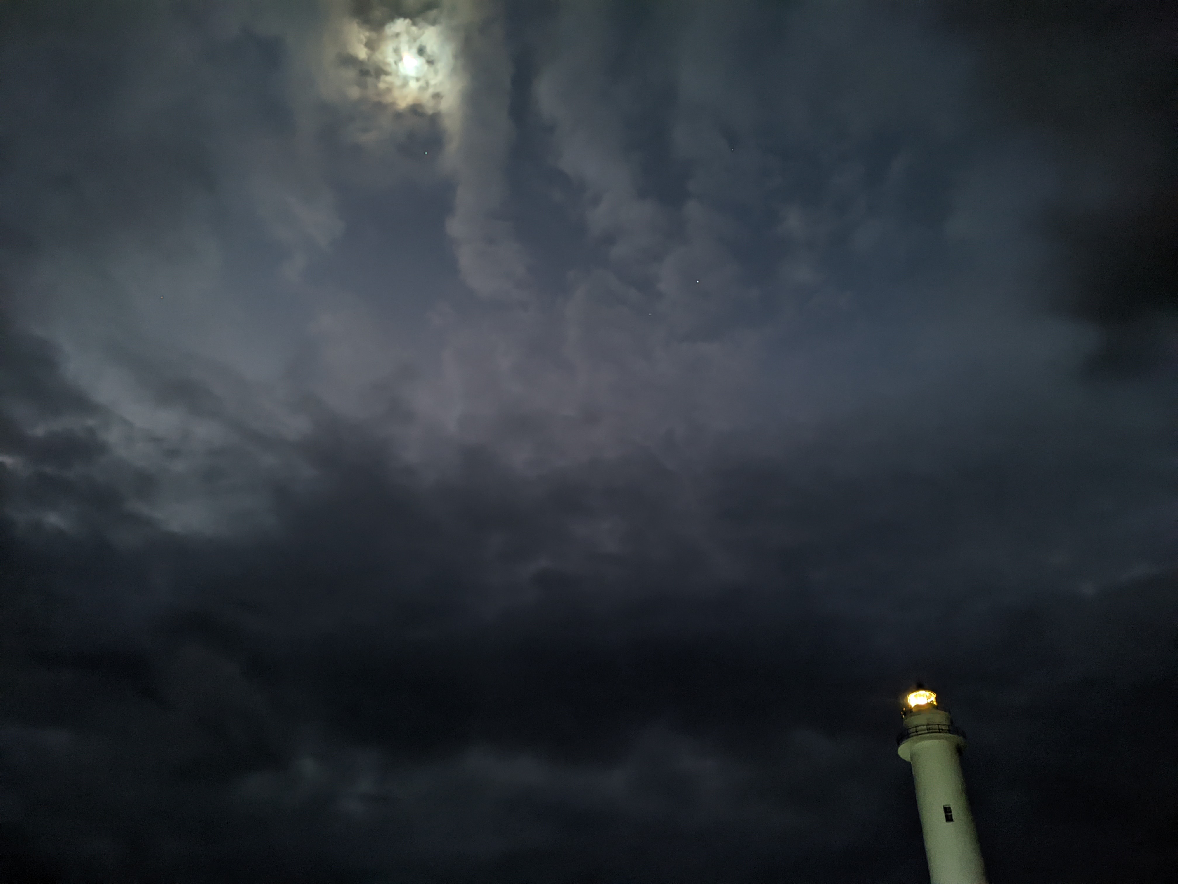 dark, cloudy sky with moon shining through, in lower right corner a white lighthouse is lit up