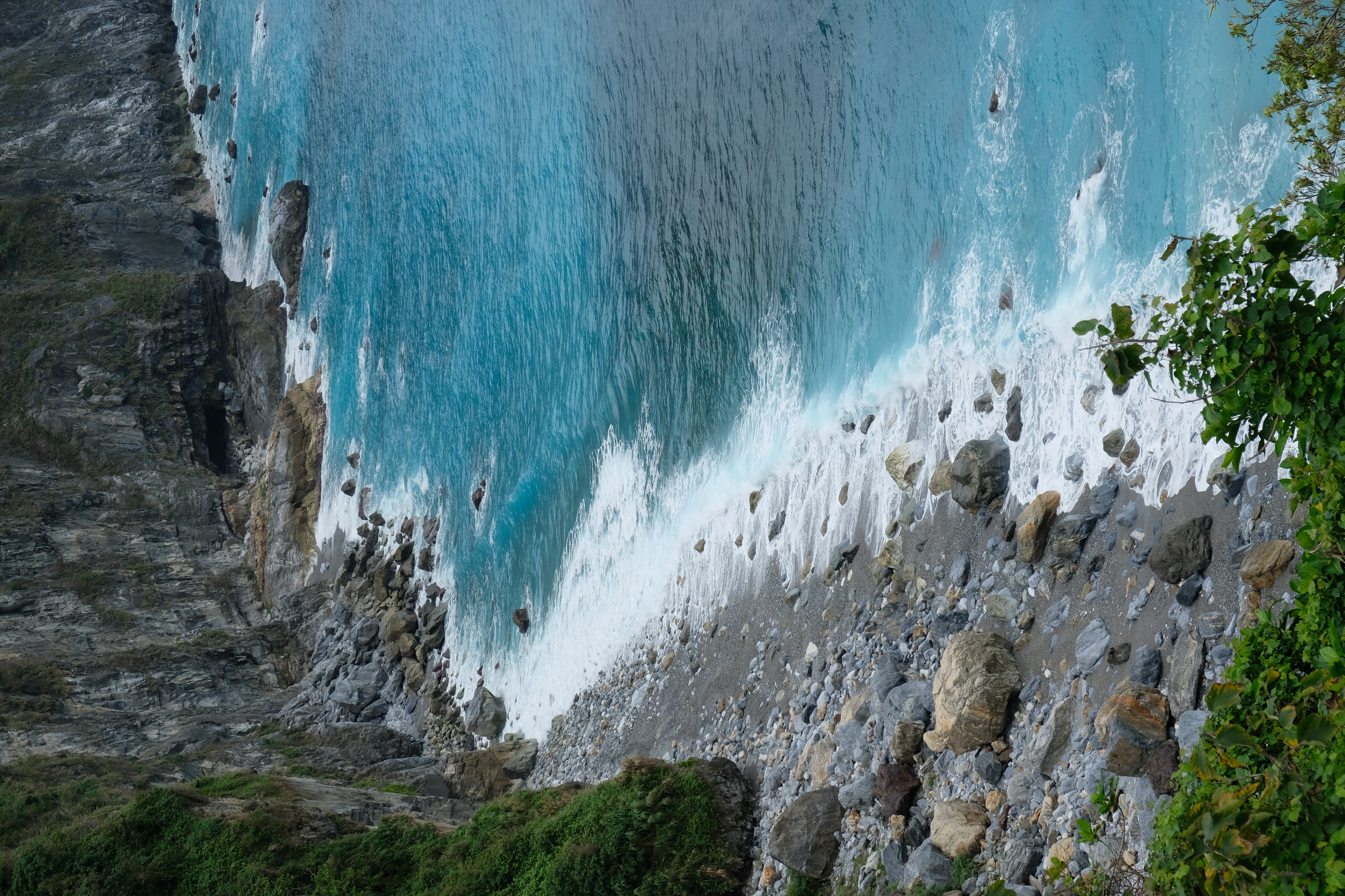 bright blue ocean with white waves crashing on a rocky beach leading up to rocky cliffs with bright green trees growing from them