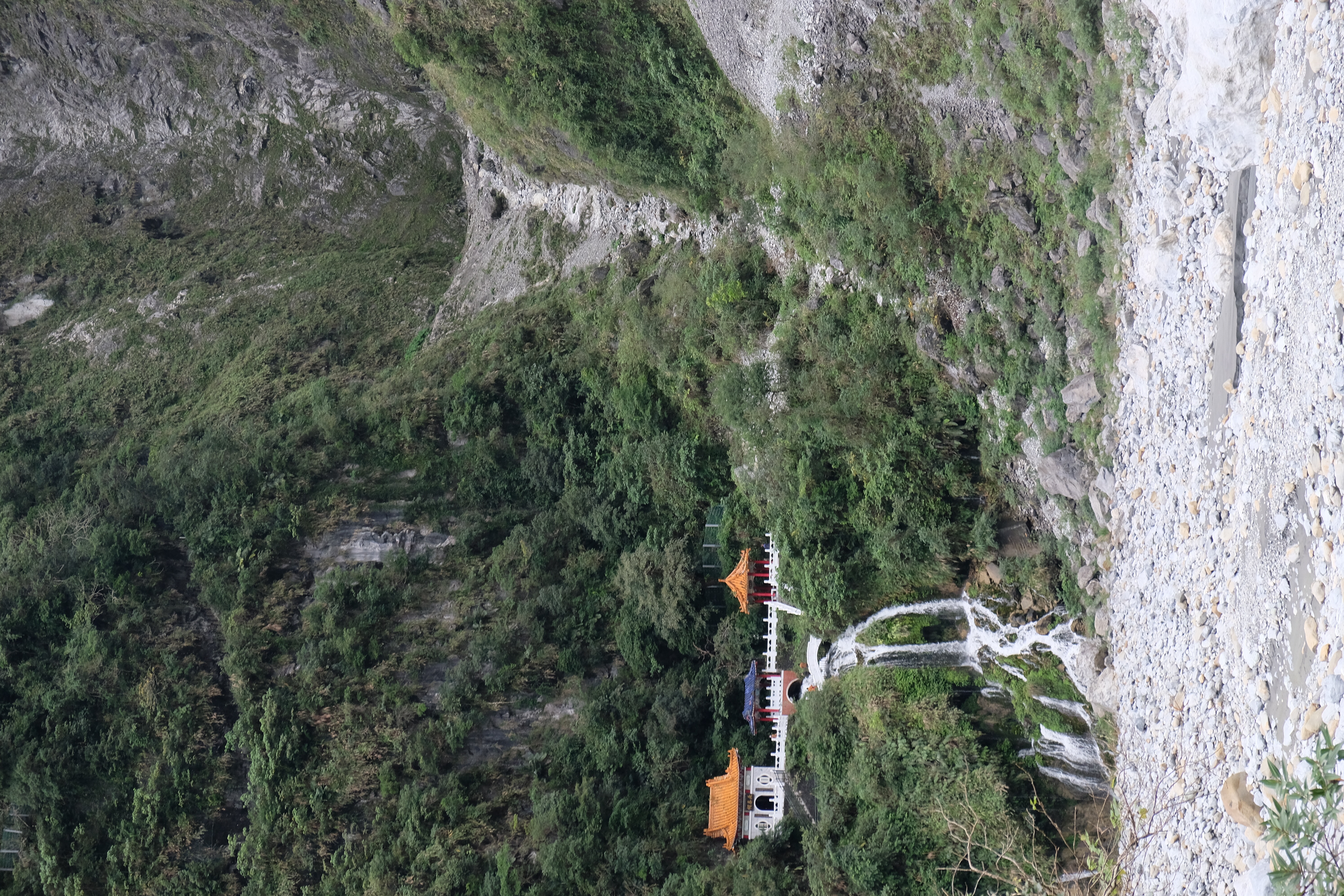 rocky, tree covered mountainside leading down to white rocky ground, about half way down the mountain on the left side is a orange, red, and white temple with a waterfall leading to the ground